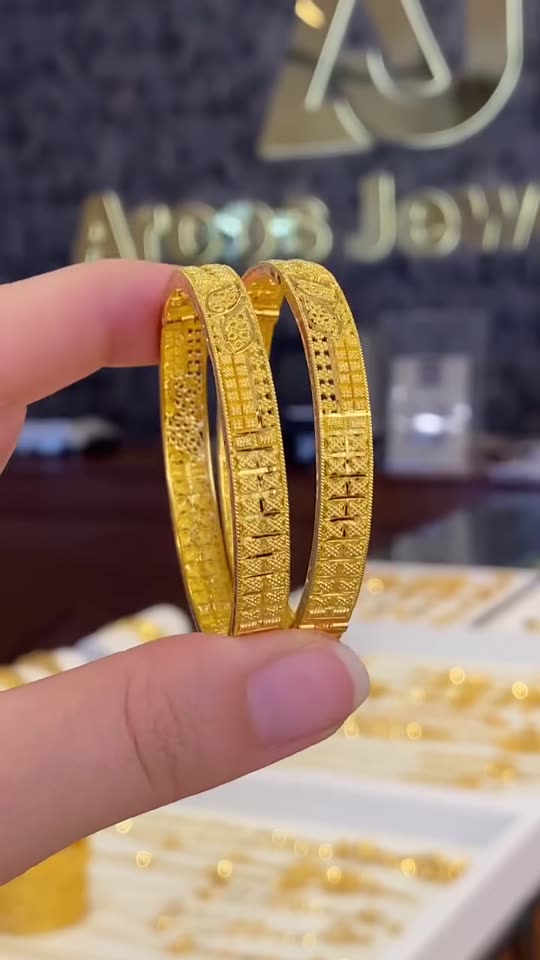 A hand holding a pair of patterned 21 carat gold bangles up to the light at the counter