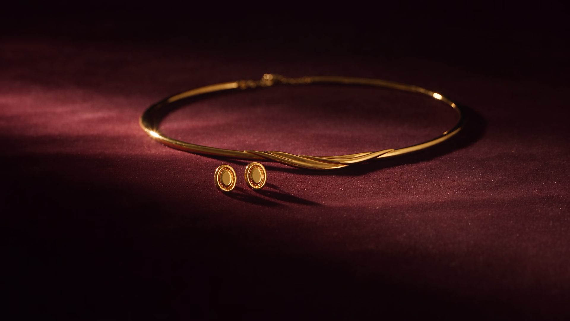 A matched gold necklace and earrings on deep maroon velvet, the camera easing in